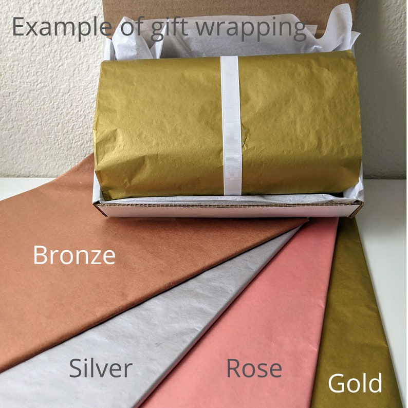 May include: A gift box wrapped in gold tissue paper and secured with a white ribbon. The box is inside a cardboard box, with tissue paper and several sheets of colored tissue paper in bronze, silver, rose, and gold.