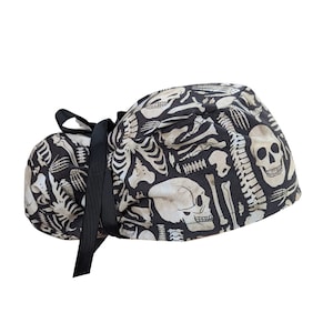 May include: A black and off-white skull and skeleton print scrub cap with a black ribbon tie. The cap features a repeating pattern of human skeletal elements, including skulls, ribs, and vertebrae. The fabric is a dark background with a light bone-colored design.