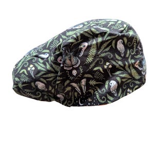 May include: A black cap with a whimsical pattern of green ferns, owls, mushrooms, moths, and skulls. The cap is made of a soft fabric and has a curved brim. The overall theme is nature and fantasy.