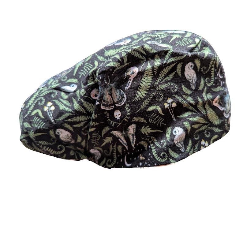 May include: A black cap with a whimsical pattern of green ferns, owls, mushrooms, moths, and skulls. The cap is made of a soft fabric and has a curved brim. The overall theme is nature and fantasy.