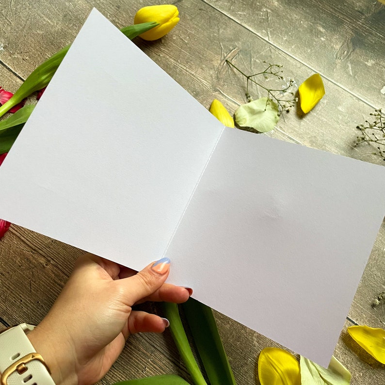 May include: A blank white card with a fold in the middle, held open by a hand. The card is surrounded by yellow and pink flowers.