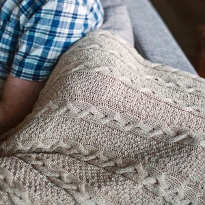 May include: A close-up of a beige cable knit throw blanket with a person wearing a blue plaid shirt in the background.