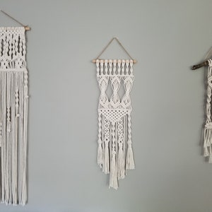 May include: Three macrame wall hangings in varying designs and sizes. The macrame pieces are made of off-white cotton cord and are suspended from wooden dowels or a tree branch. The background is a solid light gray wall.