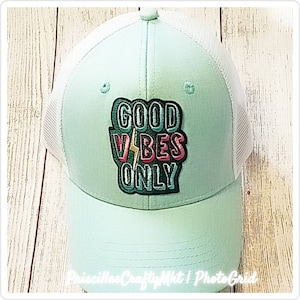 May include: A light green trucker hat with a white mesh back. The front of the hat features the embroidered phrase "GOOD VIBES ONLY" in a colorful, outlined design with a lightning bolt. The hat has a curved brim.
