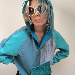 Può includere: Una felpa con cappuccio turchese e blu con frange e una tasca frontale. La felpa ha un design unico con tessuto strutturato sulle maniche e sul cappuccio. La persona indossa occhiali da sole color crema.