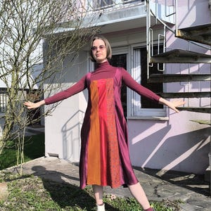 Puede incluir: Un vestido burdeos con un diseño de bloques de color con paneles rojos y naranjas. El vestido se usa sobre un cuello alto burdeos y se combina con botines blancos. El fondo incluye una casa, una escalera de caracol y un árbol.