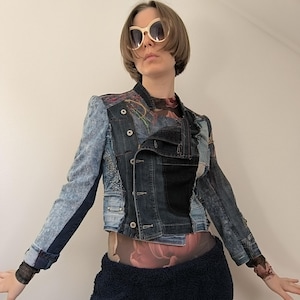 May include: A blue denim jacket with distressed details and a floral print lining. The jacket has a button closure and a collar. The model is wearing the jacket with a black skirt and white sunglasses.