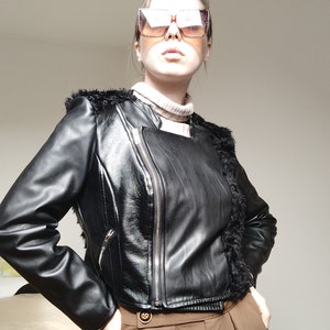 Puede incluir: Una chaqueta negra de cuero sintético con cierre de cremallera y ribete de piel en los hombros. La persona lleva pantalones marrones y gafas de sol cuadradas grandes. El atuendo es una mezcla de texturas y estilos.