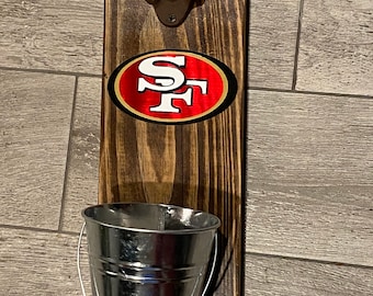 Puede incluir: Abrebotellas de madera con un abrebotellas de metal y un pequeño cubo de metal debajo. El abrebotellas tiene un logotipo de los San Francisco 49ers.