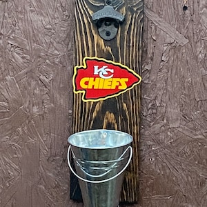 May include: A wooden bottle opener with a metal bottle opener attached. The bottle opener has a small metal bucket attached to the bottom. The bottle opener has a Kansas City Chiefs logo on it.