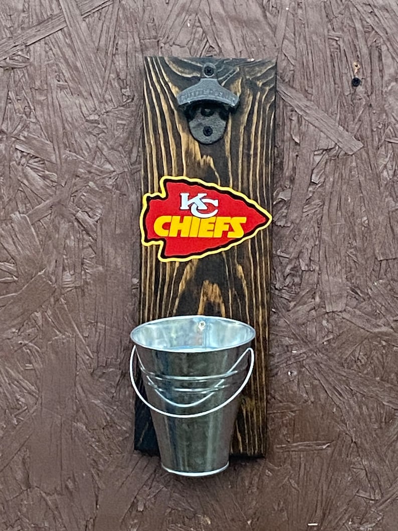 May include: A wooden bottle opener with a metal bottle opener attached. The bottle opener has a small metal bucket attached to the bottom. The bottle opener has a Kansas City Chiefs logo on it.