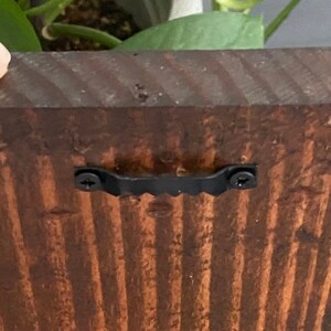 May include: A close-up of a dark brown wooden sign with a black metal hanging bracket attached to the back. The bracket has two screws visible.