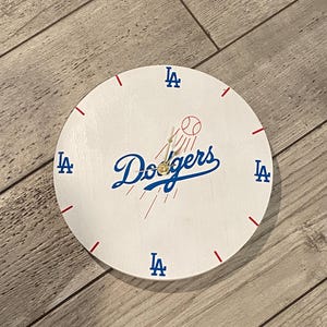 May include: White round clock featuring the Los Angeles Dodgers logo in blue script with a baseball graphic. The clock has red dashes for hour markers and blue "LA" logos at the 12, 3, 6, and 9 o'clock positions.
