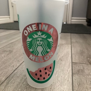 May include: A clear plastic reusable cup with a pink and green watermelon design. The cup has a sticker with the text "One in a Melon" and a green Starbucks logo.