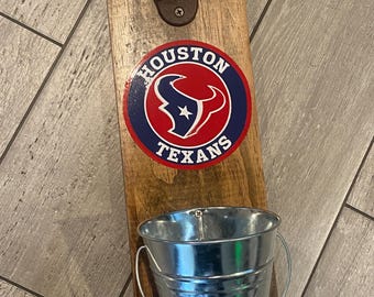 May include: Wooden bottle opener with a metal bottle opener and a small metal bucket attached. The bottle opener features the Houston Texans logo.