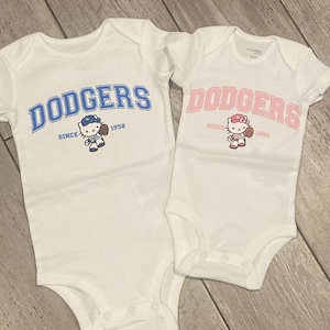 May include: Two white baby bodysuits with the word "Dodgers" printed in blue and pink on each. The bodysuits feature a cartoon character wearing a baseball cap and holding a baseball.