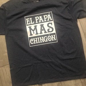 May include: A dark blue t-shirt with white text that reads "El Papá Más Chingón" in a bold, blocky font.