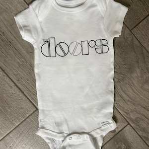 Op de afbeelding: Witte baby romper met zwarte tekst die "The Doors" in een gestileerd lettertype leest.