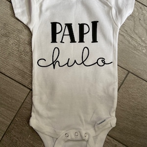 May include: White baby onesie with black text that reads "Papi chulo".
