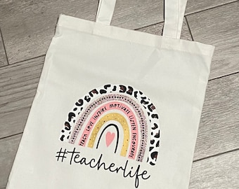 Può includere: Borsa tote bianca con un design a forma di arcobaleno e il testo "#teacherlife". L'arcobaleno ha le parole "teach, love, inspire, motivate, listen, encourage" scritte intorno ad esso.