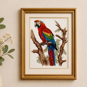 May include: A framed cross-stitch embroidery depicting a vibrant scarlet macaw perched on a brown tree branch. The bird features bright red, yellow, and blue plumage. The artwork is enclosed in a classic gold-toned frame with a white mat border.
