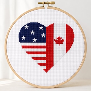 May include: A circular wooden embroidery hoop containing a cross-stitch design of a heart. The left half features the American flag with white stars on a blue field and red and white stripes. The right half features the Canadian flag with a red maple leaf.