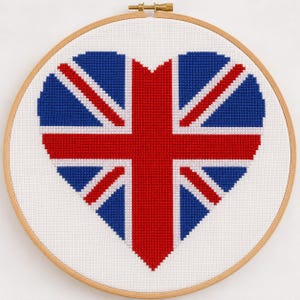 May include: A circular wooden embroidery hoop containing white fabric with a cross-stitch design of a heart shaped like the Union Jack flag. The pattern features red, white, and blue thread forming the iconic British flag design on a white background.