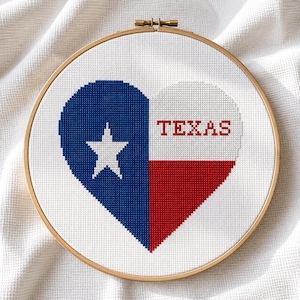 Puede incluir: Un bastidor de bordado circular de madera que contiene un diseño de punto de cruz en forma de corazón que representa la bandera del estado de Texas. El lado izquierdo es azul con una estrella blanca y el derecho tiene franjas horizontales blancas y rojas con la palabra TEXAS.