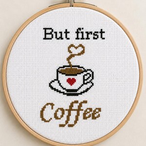 May include: A circular wooden embroidery hoop containing white Aida fabric with a cross-stitch design. The text reads But first in black and Coffee in brown. A coffee cup with a red heart and brown steam shaped like a heart is centred.