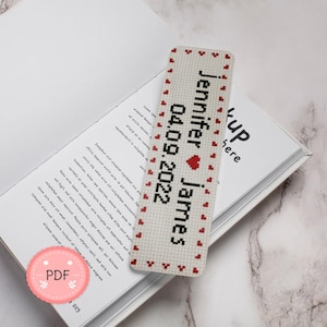 May include: A personalized, cross-stitched bookmark with the names "Jennifer" and "James" and the date "04.09.2022" in black. Red hearts border the edges. The bookmark rests on an open book.