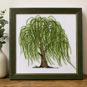 May include: A framed cross-stitch embroidery depicting a weeping willow tree with vibrant green foliage and a brown trunk, set against a white grid fabric background. The piece is enclosed in a dark green wooden frame with a thin gold inner border.