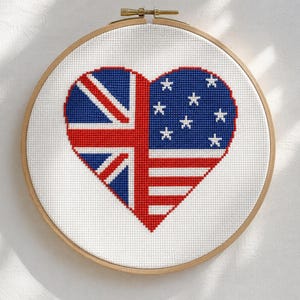 May include: A circular wooden embroidery hoop containing a cross-stitch pattern of a heart shape. The left side of the heart features the Union Jack flag design, while the right side displays the stars and stripes of the United States flag.