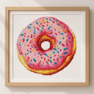 May include: A cross-stitch embroidery of a round donut with pink icing and multicolored sprinkles, featuring a golden-brown fried dough base, centered within a square light-colored wooden frame against a plain white background.