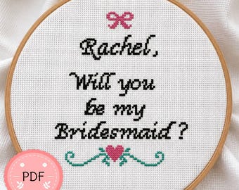 May include: A circular wooden embroidery hoop containing white Aida fabric with cross-stitched text that reads Rachel, Will you be my Bridesmaid? The design features a small pink bow at the top and a pink heart with green flourishes at the bottom.