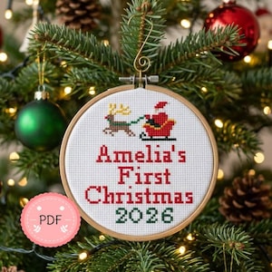 Pode incluir: Um enfeite de Natal em ponto cruz num aro de madeira, com o Pai Natal num trenó puxado por uma rena. O texto diz "Amelia's First Christmas 2026" em vermelho. O enfeite está pendurado numa árvore de Natal.