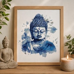 May include: A framed cross-stitch embroidery featuring a blue watercolor-style portrait of a Buddha figure with closed eyes and a serene expression, set against a white fabric background within a light-colored wooden frame.
