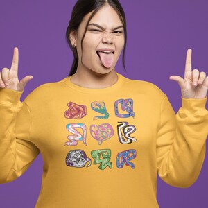 Pode incluir: Uma mulher vestindo uma jaqueta de moletom amarela com um design de cobra colorida. Ela está fazendo um gesto de rock and roll com ambas as mãos.