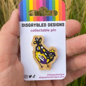 Può includere: Uno spillo in legno con un uccello stilizzato nero, viola e giallo con accenti bianchi. Lo spillo è su una carta con il testo "Disgaybled Designs" e "collectable pin".