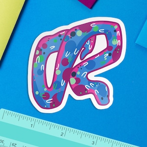 May include: A sticker featuring an abstract design in pink and blue, shaped like the letter "R".
