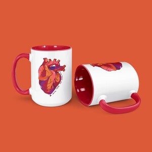 以下が含まれることがあります： 赤い内側の白いセラミック製のマグカップが2つ。1つのマグカップには、人間の心臓のカラフルなイラストと「You make my heart happy」というテキストが描かれています。
