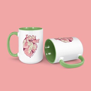 以下が含まれることがあります： 緑色の縁取りが付いた白いセラミック製のマグカップが2つ。1つのマグカップには、ピンクの解剖学的心臓と「You make my heart beat」というピンクの文字が書かれています。もう1つのマグカップは空です。
