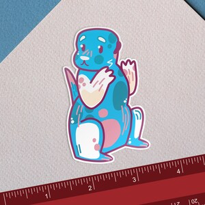 Op de afbeelding: Een blauwe en roze cartoon-otter sticker met witte vlekken en een witte poot. De otter zwaait met zijn rechterpoot.