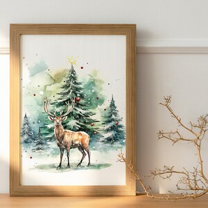 以下が含まれることがあります： 雪の降る森にいる鹿を描いた、額入りの水彩画。クリスマスツリーが描かれています。フレームは明るい茶色の木製です。赤いオーナメントと星が木を飾っています。全体的な配色は緑、茶色、白です。
