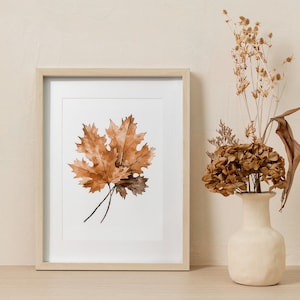 May include: A framed print of a watercolour painting of three brown maple leaves on a white background. The frame is light wood and the print is hanging on a beige wall.