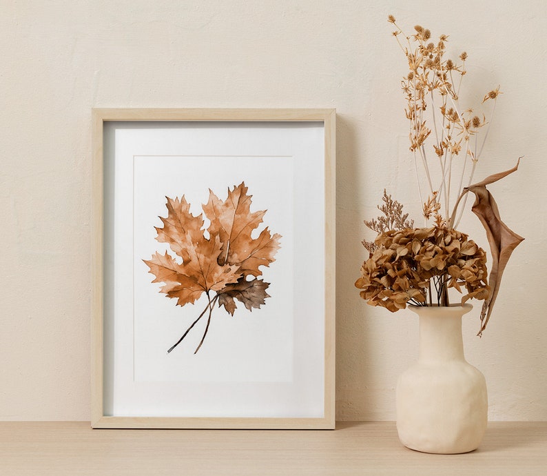 May include: A framed print of a watercolour painting of three brown maple leaves on a white background. The frame is light wood and the print is hanging on a beige wall.