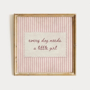 Puede incluir: Un cuadro cuadrado con marco de metal dorado y fondo de rayas verticales en rosa claro y blanco. En el centro, un papel rectangular de color crema con bordes festoneados muestra el texto en rojo oscuro: every dog needs a little girl.