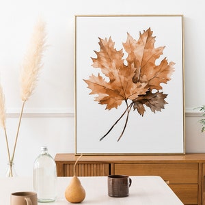 May include: A watercolour painting of a brown maple leaf in a gold frame, hanging on a white wall above a wooden dresser.