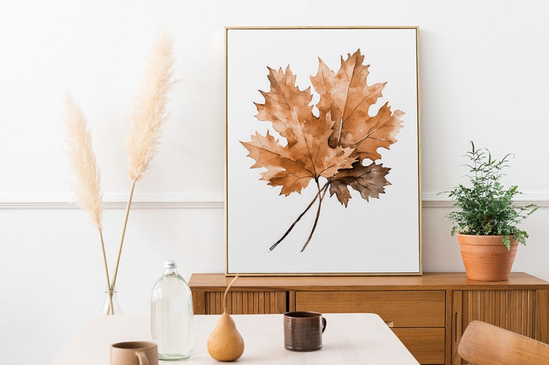 May include: A watercolour painting of a brown maple leaf in a gold frame, hanging on a white wall above a wooden dresser.