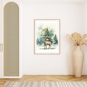 以下が含まれることがあります： 雪の降る森にいる鹿の額装された水彩画。アートワークは明るい木製のフレームに入っています。シーンは、木のてっぺんに星がある冬の風景です。