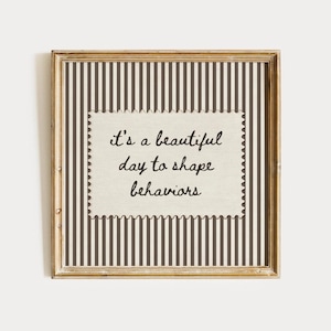 May include: A square framed wall art piece featuring a cream-coloured centre with a scalloped edge, printed with the black cursive text 'it's a beautiful day to shape behaviours'. The background consists of vertical dark brown and white stripes.